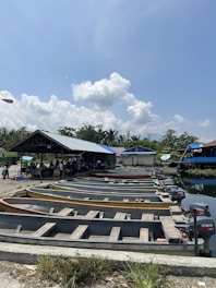 A row of small wooden boats with outboard motors is aligned on a concrete ramp by the water's edge. The area is shaded by a large roofed structure where a group of people is gathered. A clear blue sky with scattered fluffy clouds forms the backdrop, and lush green trees are visible in the background.