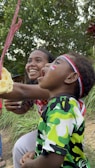 A child and an adult are participating in a fun outdoor activity, where the child is attempting to catch a snack hanging from a string. The child is wearing a colorful camouflage-patterned shirt and a red, white, and blue headband with a small flag painted on the cheek. The adult, smiling broadly, is encouraging the child. The scene is set in a grassy area with green foliage in the background, creating a natural and lively atmosphere.