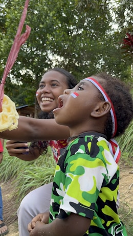 A child and an adult are participating in a fun outdoor activity, where the child is attempting to catch a snack hanging from a string. The child is wearing a colorful camouflage-patterned shirt and a red, white, and blue headband with a small flag painted on the cheek. The adult, smiling broadly, is encouraging the child. The scene is set in a grassy area with green foliage in the background, creating a natural and lively atmosphere.