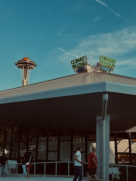 A modern building with 'Climate Pledge Arena' signage is in the foreground. In the background, a distinctive tower with an orange top can be seen against a blue sky. Several people are walking on the ground level, near the building, creating a lively atmosphere.