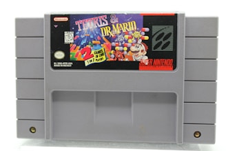 A gray Super Nintendo Entertainment System (SNES) cartridge displaying the label for 'Tetris & Dr. Mario'. The label features colorful graphics, including falling blocks from Tetris and Dr. Mario characters like Mario, viruses, and capsules. It also includes the Nintendo logo and information indicating it's a game for one or two players.