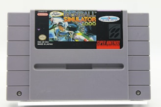A Super Nintendo Entertainment System (SNES) game cartridge for 'Baseball Simulator 1.000' by Culture Brain. The cartridge is gray with a label featuring a vibrant illustration of baseball players in action, displaying dynamic poses and bright colors. The label includes logos for Nintendo and Culture Brain, as well as text indicating the game title and region as 'Made in Japan.'