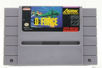 A gray Super Nintendo Entertainment System (SNES) cartridge featuring the cover art for the game 'D-Force'. The label includes a helicopter against a blue sky background, and the logos for Nintendo and Asmik Corporation of America.