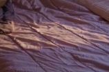 A close-up view of a bed with crumpled, lavender-colored sheets. Soft lighting casts gentle shadows and highlights on the fabric, suggesting an early morning or late afternoon ambiance. A portion of a pillow can be seen on the left side, and another pillow is partially visible on the right side.