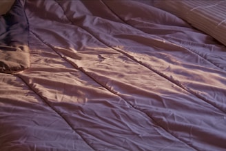 A close-up view of a bed with crumpled, lavender-colored sheets. Soft lighting casts gentle shadows and highlights on the fabric, suggesting an early morning or late afternoon ambiance. A portion of a pillow can be seen on the left side, and another pillow is partially visible on the right side.
