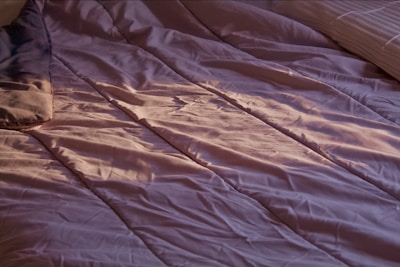 A close-up view of a bed with crumpled, lavender-colored sheets. Soft lighting casts gentle shadows and highlights on the fabric, suggesting an early morning or late afternoon ambiance. A portion of a pillow can be seen on the left side, and another pillow is partially visible on the right side.