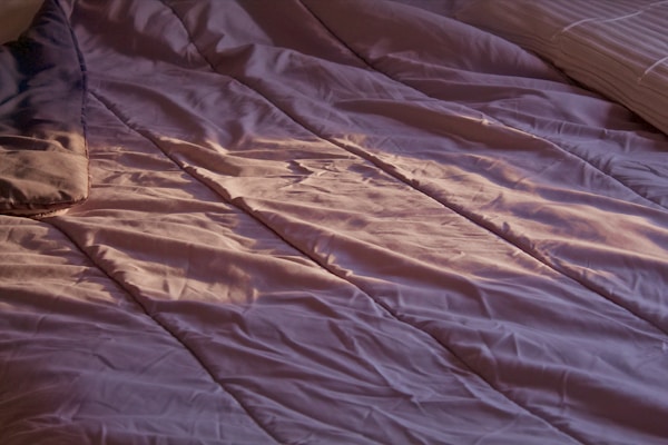 A close-up view of a bed with crumpled, lavender-colored sheets. Soft lighting casts gentle shadows and highlights on the fabric, suggesting an early morning or late afternoon ambiance. A portion of a pillow can be seen on the left side, and another pillow is partially visible on the right side.
