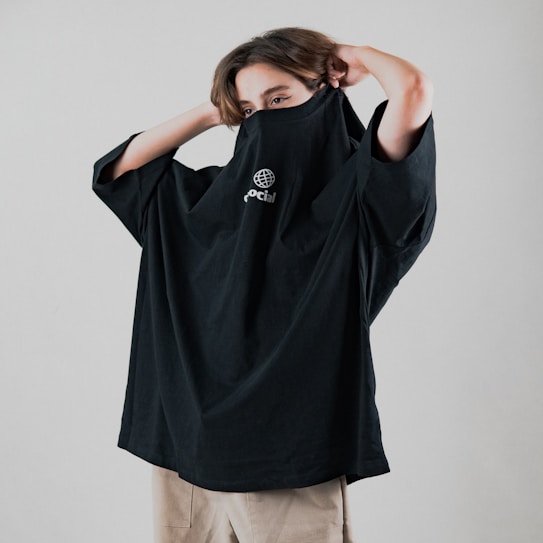 A person is pulling a black oversized shirt over their face, revealing only their eyes. The shirt features a white graphic logo and the word 'social'. The person has light brown hair and is standing against a neutral background, wearing light-colored pants.