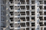 High-rise apartment building with multiple floors and balconies. The facade features a mix of white and grey tones with glass railings. Some balconies have sacks or construction materials visible, indicating ongoing construction or maintenance work.