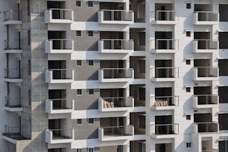 High-rise apartment building with multiple floors and balconies. The facade features a mix of white and grey tones with glass railings. Some balconies have sacks or construction materials visible, indicating ongoing construction or maintenance work.