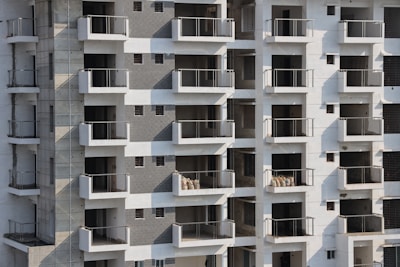 High-rise apartment building with multiple floors and balconies. The facade features a mix of white and grey tones with glass railings. Some balconies have sacks or construction materials visible, indicating ongoing construction or maintenance work.