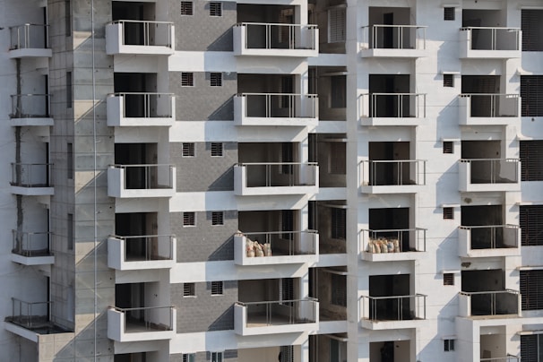 High-rise apartment building with multiple floors and balconies. The facade features a mix of white and grey tones with glass railings. Some balconies have sacks or construction materials visible, indicating ongoing construction or maintenance work.