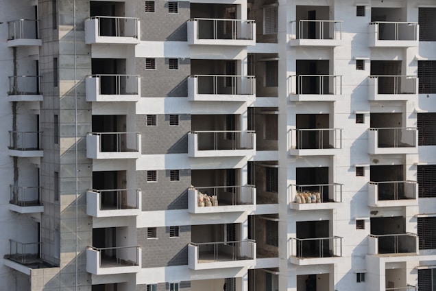 High-rise apartment building with multiple floors and balconies. The facade features a mix of white and grey tones with glass railings. Some balconies have sacks or construction materials visible, indicating ongoing construction or maintenance work.