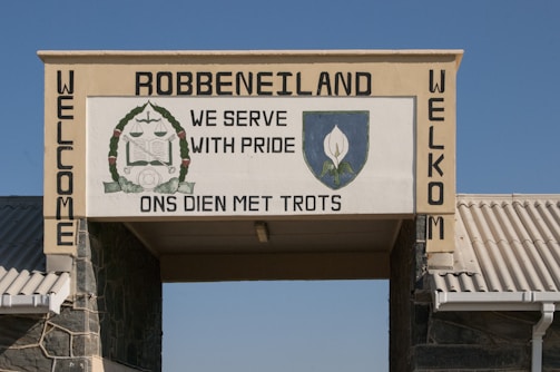 A stone archway with a sign displays the name Robben Island and phrases 'We Serve With Pride' and 'Ons Dien Met Trots'. The sign features a crest with symbolic imagery.