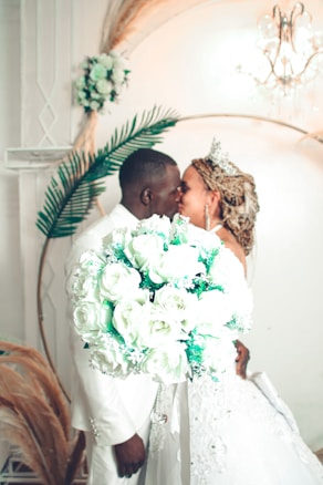 A couple dressed in elegant wedding attire is sharing a kiss. The bride is holding a beautiful bouquet of white roses with green accents. In the background, there is a decorative setting including a chandelier and large green leaves, adding to the romantic ambiance.