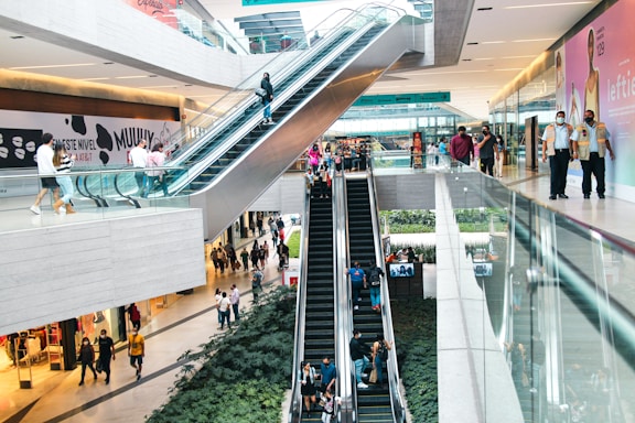 A modern shopping mall features multiple levels connected by escalators. Shoppers are casually walking on different floors, and some are using the escalators. The mall is spacious, with large advertisements on the walls and a mix of glass and metallic architecture. Green plants are situated near the escalators, adding a touch of nature to the contemporary setting.
