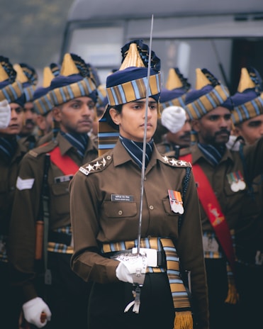 A group of uniformed individuals, likely military personnel, are standing in formation. The focus is on one person in the foreground who is holding a sword and wearing a decorated uniform with medals on the chest. Their hats are uniquely styled with blue and golden stripes.