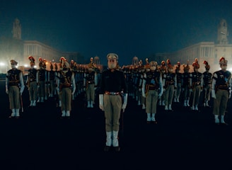 A group of uniformed personnel stands in a symmetrical formation on a dimly lit, open area. Each person wears a khaki uniform with a distinctive red plume on their hats. The surrounding environment is shrouded in a misty atmosphere with large architectural buildings visible in the background.
