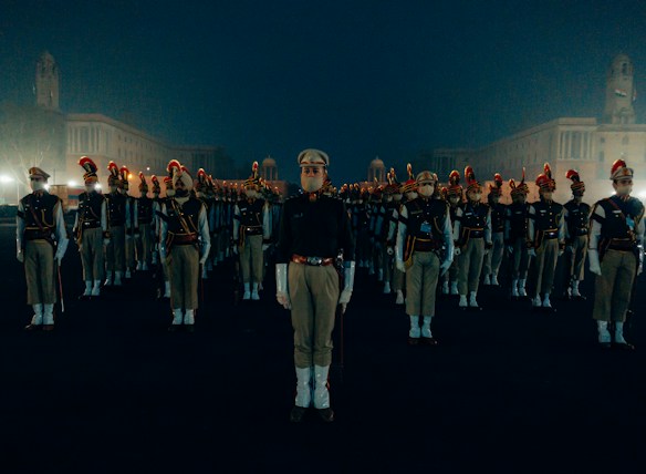 A group of uniformed personnel stands in a symmetrical formation on a dimly lit, open area. Each person wears a khaki uniform with a distinctive red plume on their hats. The surrounding environment is shrouded in a misty atmosphere with large architectural buildings visible in the background.