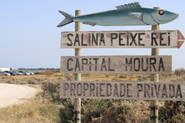 A signpost with a blue fish on top points in a direction, featuring three wooden planks with the inscriptions 'SALINA PEIXE REI,' 'CAPITAL MOURA,' and 'PROPRIEDADE PRIVADA.' In the background, a dirt road leads to a row of parked cars and a flat landscape with sparse vegetation under a clear sky.