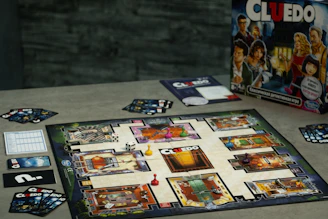 A board game setup of Cluedo is displayed on a table. The game board showcases various rooms depicted in a mansion layout, with colored tokens placed on certain locations. Several Cluedo cards and dice are arranged around the board. The game box, featuring illustrated characters, is positioned upright in the background.