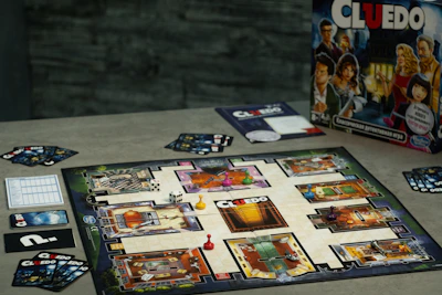 A board game setup of Cluedo is displayed on a table. The game board showcases various rooms depicted in a mansion layout, with colored tokens placed on certain locations. Several Cluedo cards and dice are arranged around the board. The game box, featuring illustrated characters, is positioned upright in the background.