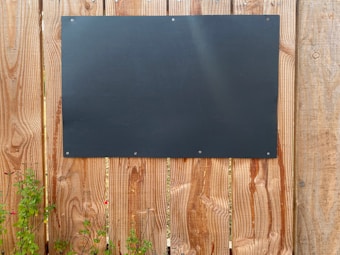 A large black rectangular board is mounted on a wooden fence. The wood has a vertical plank design with a natural grain pattern. Small green plants with red flowers are growing at the base of the fence.