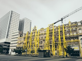 A historic building is under renovation with bright yellow scaffolding on the facade. The building is set against a backdrop of modern high-rise structures. A crane is visible above, indicating ongoing construction work in the area.