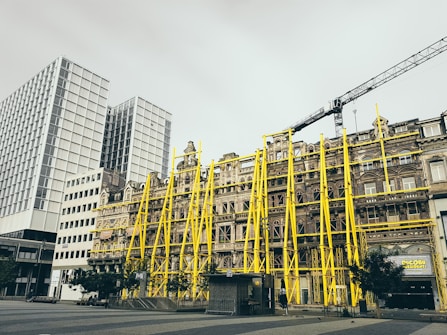 A historic building is under renovation with bright yellow scaffolding on the facade. The building is set against a backdrop of modern high-rise structures. A crane is visible above, indicating ongoing construction work in the area.
