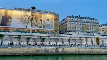 An urban riverside scene with people sitting along the embankment. A large building features a prominent advertisement with fashion images. In the background, there are classic European-style buildings with trees lining the street.