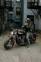 A person wearing a black helmet and a dark jacket is seated on a black motorcycle in an urban alleyway. The motorcycle has a prominent 'Indian' logo on its side. The background features brick walls and fire escapes, giving an industrial vibe.