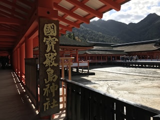 A traditional Japanese shrine with red wooden structures, featuring a large sign with Japanese characters. The architecture includes long corridors and elevated wooden decking with a mountainous backdrop. A group of people can be seen walking in the courtyard area.