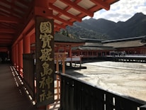 A traditional Japanese shrine with red wooden structures, featuring a large sign with Japanese characters. The architecture includes long corridors and elevated wooden decking with a mountainous backdrop. A group of people can be seen walking in the courtyard area.