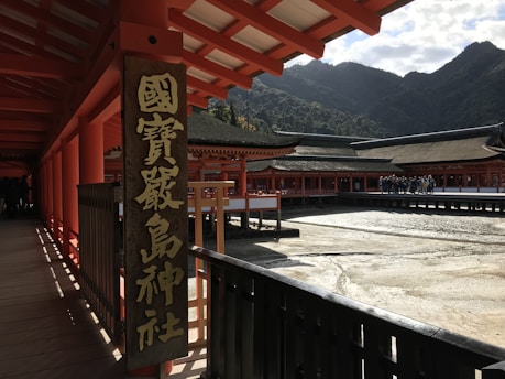 A traditional Japanese shrine with red wooden structures, featuring a large sign with Japanese characters. The architecture includes long corridors and elevated wooden decking with a mountainous backdrop. A group of people can be seen walking in the courtyard area.