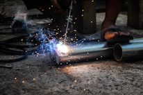 A person is engaged in metal welding using a torch, with bright sparks and smoke emanating from the point of contact on two metal pipes lying on a concrete floor. The feet of the person in sandals are visible, highlighting a focus on the welding task in a workshop or industrial setting.