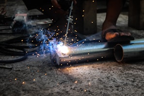 A person is engaged in metal welding using a torch, with bright sparks and smoke emanating from the point of contact on two metal pipes lying on a concrete floor. The feet of the person in sandals are visible, highlighting a focus on the welding task in a workshop or industrial setting.