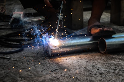 A person is engaged in metal welding using a torch, with bright sparks and smoke emanating from the point of contact on two metal pipes lying on a concrete floor. The feet of the person in sandals are visible, highlighting a focus on the welding task in a workshop or industrial setting.