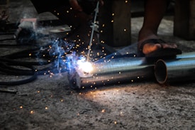 A person is engaged in metal welding using a torch, with bright sparks and smoke emanating from the point of contact on two metal pipes lying on a concrete floor. The feet of the person in sandals are visible, highlighting a focus on the welding task in a workshop or industrial setting.