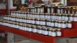 A neatly arranged display of various jars set on red wooden shelves, featuring honey and other preserves. Each jar has a label with white text, and the setting suggests a rustic or farm-style market. The background includes boxes and decorations, adding to the cozy atmosphere.