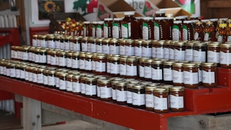 A neatly arranged display of various jars set on red wooden shelves, featuring honey and other preserves. Each jar has a label with white text, and the setting suggests a rustic or farm-style market. The background includes boxes and decorations, adding to the cozy atmosphere.