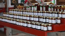 A neatly arranged display of various jars set on red wooden shelves, featuring honey and other preserves. Each jar has a label with white text, and the setting suggests a rustic or farm-style market. The background includes boxes and decorations, adding to the cozy atmosphere.