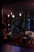 A dimly lit room features modern furniture, including curved chairs and a large, reflective Rotalux light reflector. The walls have vertical slits creating patterns with the light. Several spherical lights hang from the ceiling, adding to the atmospheric and stylish ambiance. There is a blend of deep reds, blues, and subtle oranges creating a cozy yet sophisticated interior space.