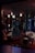 A dimly lit room features modern furniture, including curved chairs and a large, reflective Rotalux light reflector. The walls have vertical slits creating patterns with the light. Several spherical lights hang from the ceiling, adding to the atmospheric and stylish ambiance. There is a blend of deep reds, blues, and subtle oranges creating a cozy yet sophisticated interior space.