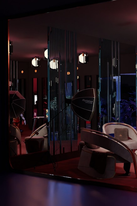 A dimly lit room features modern furniture, including curved chairs and a large, reflective Rotalux light reflector. The walls have vertical slits creating patterns with the light. Several spherical lights hang from the ceiling, adding to the atmospheric and stylish ambiance. There is a blend of deep reds, blues, and subtle oranges creating a cozy yet sophisticated interior space.