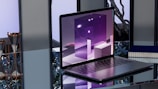 A sleek, modern laptop placed on a reflective glass surface. The screen displays abstract, geometric shapes in shades of purple with bright highlights. To the left, two hourglasses with gold frames are seen. In the background, blurred objects and a potted plant provide an industrial yet sophisticated ambiance.