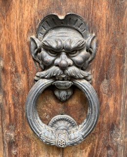 An intricately designed metal door knocker is mounted on a wooden door. The knocker features a detailed face with exaggerated features including a bushy mustache, sharp eyes, and pointed ears. The ring is held in the mouth of the face and appears ornate with carved patterns.