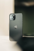 A sleek, modern smartphone with a metallic finish is standing upright on a flat surface. The device features three camera lenses within a rectangular module positioned in the top left corner. The iconic apple logo is prominently displayed at the center of the back of the phone.