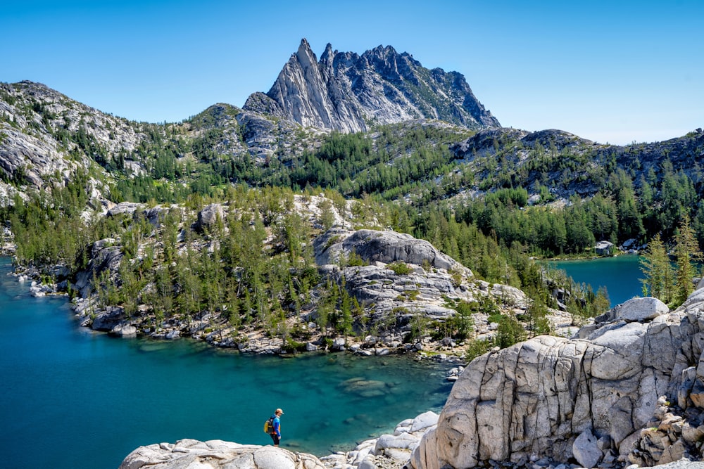 a person standing on a rocky shore by a lake and mountains