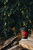 A black disposable coffee cup with a red logo and the words 'marrone rosso' printed on it sits on a stack of rustic wooden logs. A black straw is inserted into the cup, and the background features lush green foliage, creating a natural and serene setting.