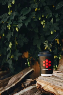 A black disposable coffee cup with a red logo and the words 'marrone rosso' printed on it sits on a stack of rustic wooden logs. A black straw is inserted into the cup, and the background features lush green foliage, creating a natural and serene setting.